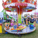 Kolorowa karuzela z figurkami dziecięcymi na tle wesołego miasteczka. Wokół dużo ludzi, kolorowy namiot i ciężarówka. Trawa pod nogami, jasno na niebie.