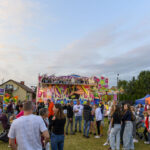 Tłum ludzi na tle wesołego miasteczka z kolorowym, różowym diabelskim młynem. Namioty w pomarańczowym i niebieskim kolorze. Słoneczne niebo z chmurami.