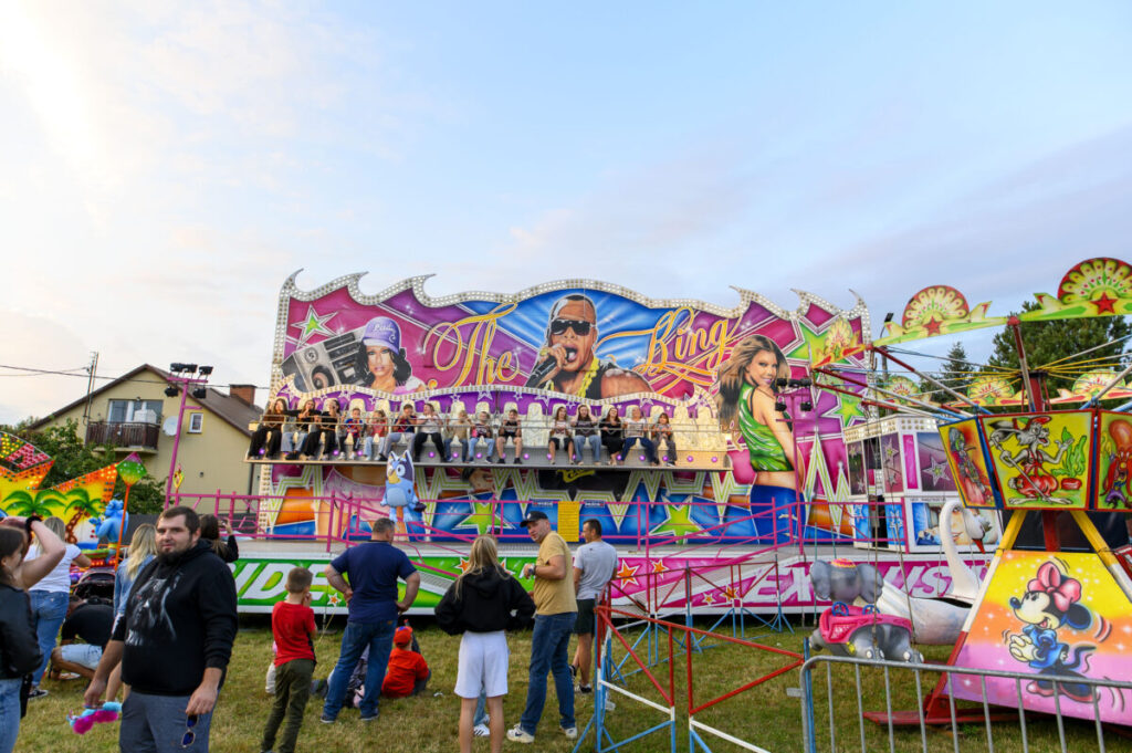 Kolorowy, wesoły lunapark z różowymi i zielonymi elementami. Na pierwszym planie karuzela, w tle tłum ludzi. Obok domy i drzewa.