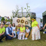 Grupa osób w kolorowych strojach siedzi i stoi na tle wieńców dożynkowych. Tło to namiot i dekoracje z kukurydzy. Ziemia jest zielona.