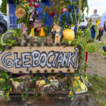 Tabliczka z napisem Głęboćanki ozdobiona kwiatami i owocami. Z tyłu ludzie na trawie. Kolory zielony i żółty.