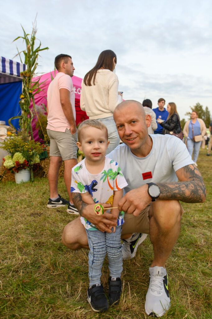 Na zdjęciu widoczny jest mężczyzna i małe dziecko na trawie. W tle ludzie i kolorowe namioty. Dziecko trzyma lizaka.
