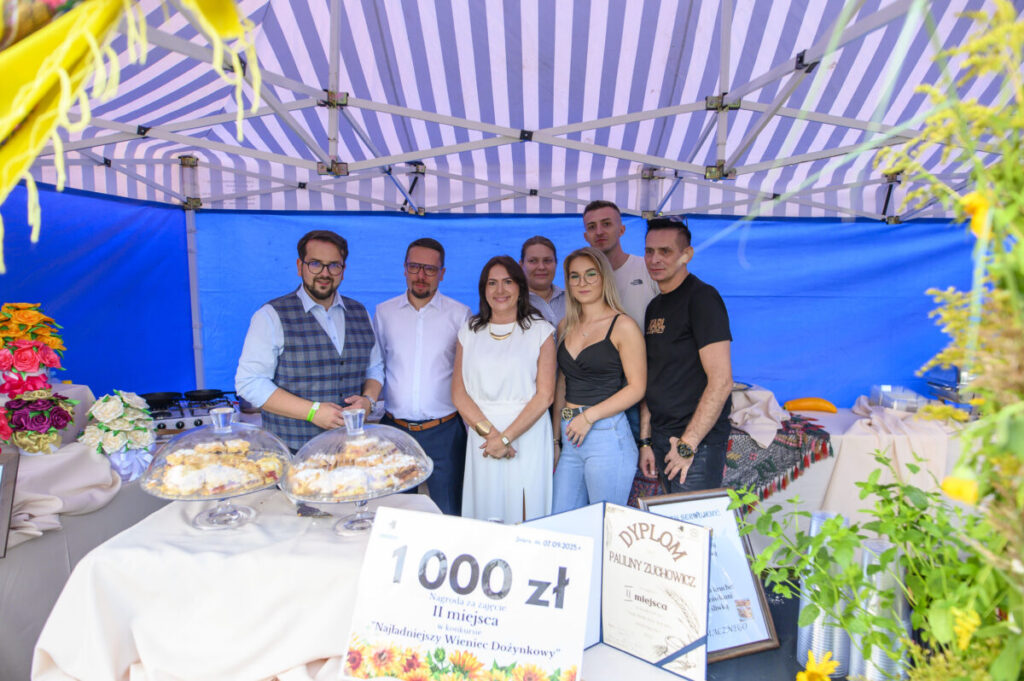 Grupa osób pod niebiesko-białym namiotem celebruje nagrody. Obok słodycze pod kloszami i kolorowe kwiaty. Tło w niebieskim odcieniu.