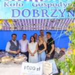Grupa ludzi stoi pod niebiesko-białym namiotem z napisem Koło Gospodyń Dobrzyn. Na stole przed nimi są ciasta i nagrody. W tle niebieskie płótno i ozdobne rośliny.