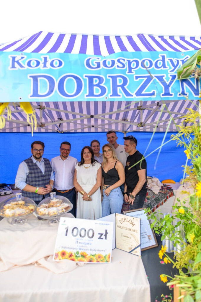Grupa ludzi stoi pod niebiesko-białym namiotem z napisem Koło Gospodyń Dobrzyn. Na stole przed nimi są ciasta i nagrody. W tle niebieskie płótno i ozdobne rośliny.