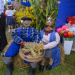 Dwa kolorowe manekiny w strojach ludowych siedzą na beli siana. W tle niebieski namiot i żółte kwiaty. Otoczenie ozdobione roślinami.