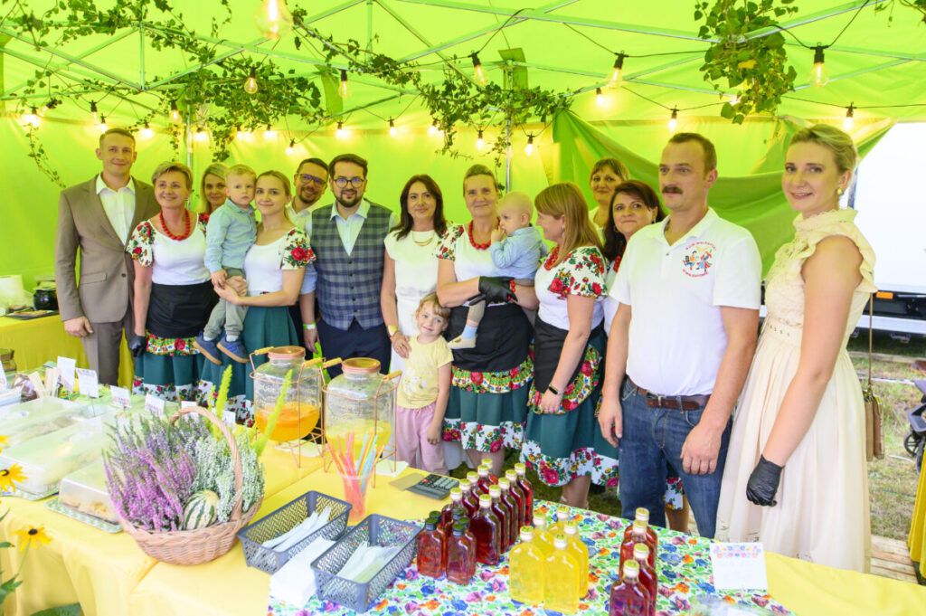 Grupa osób w kolorowych strojach stoi pod zielonym namiotem. Na stole znajdują się słoje z napojami i koszyk z lawendą. W tle girlandy i rośliny.