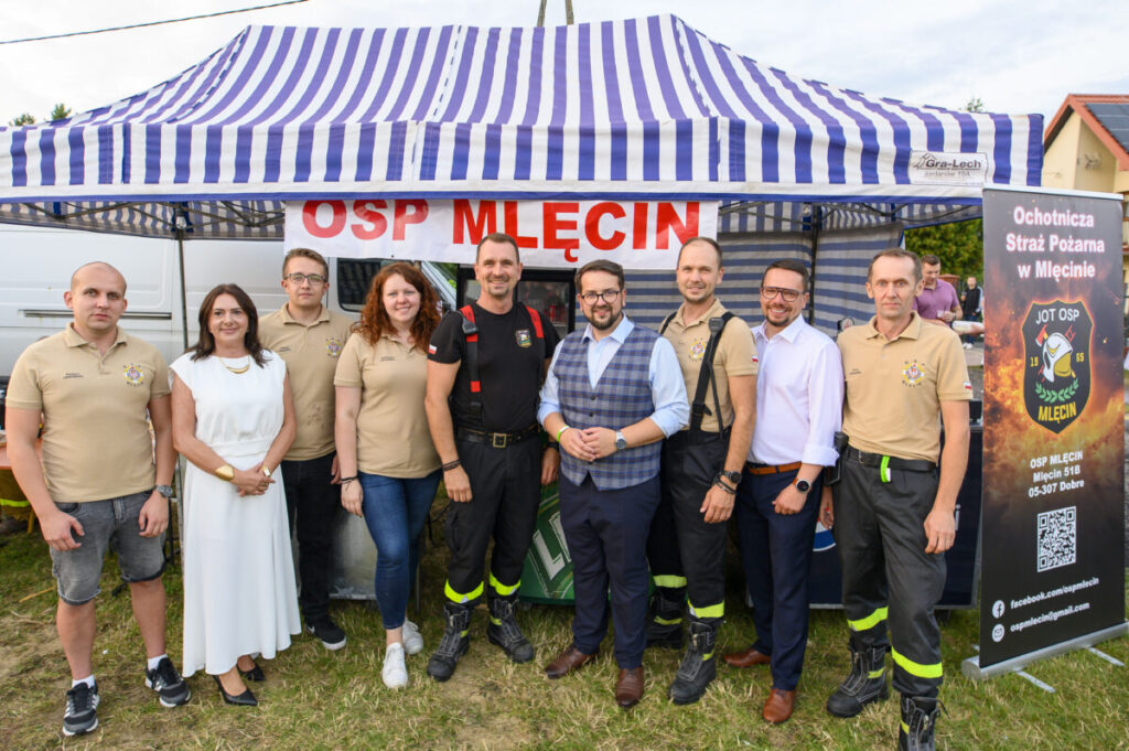 Grupa osób stoi przed niebiesko-białym namiotem z napisem OSP Mlecin. W tle widoczny jest plakat Ochotniczej Straży Pożarnej. Ludzie ubrani są w beżowe koszulki i ciemne spodnie.