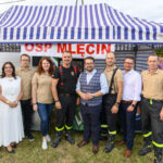 Grupa osób stoi przed niebiesko-białym namiotem z napisem OSP Mlecin. W tle widoczny jest plakat Ochotniczej Straży Pożarnej. Ludzie ubrani są w beżowe koszulki i ciemne spodnie.