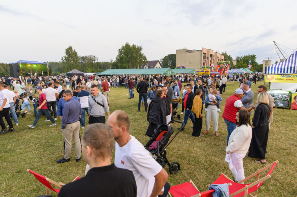Na zdjęciu widać tłum ludzi na otwartym terenie z zieloną trawą. W tle znajdują się budynki, namioty i scena z zielonym oświetleniem. Atmosfera jest festiwalowa, z kolorowymi stoiskami i rozrywkami.
