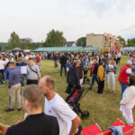 Na zdjęciu widać tłum ludzi na otwartym terenie z zieloną trawą. W tle znajdują się budynki, namioty i scena z zielonym oświetleniem. Atmosfera jest festiwalowa, z kolorowymi stoiskami i rozrywkami.