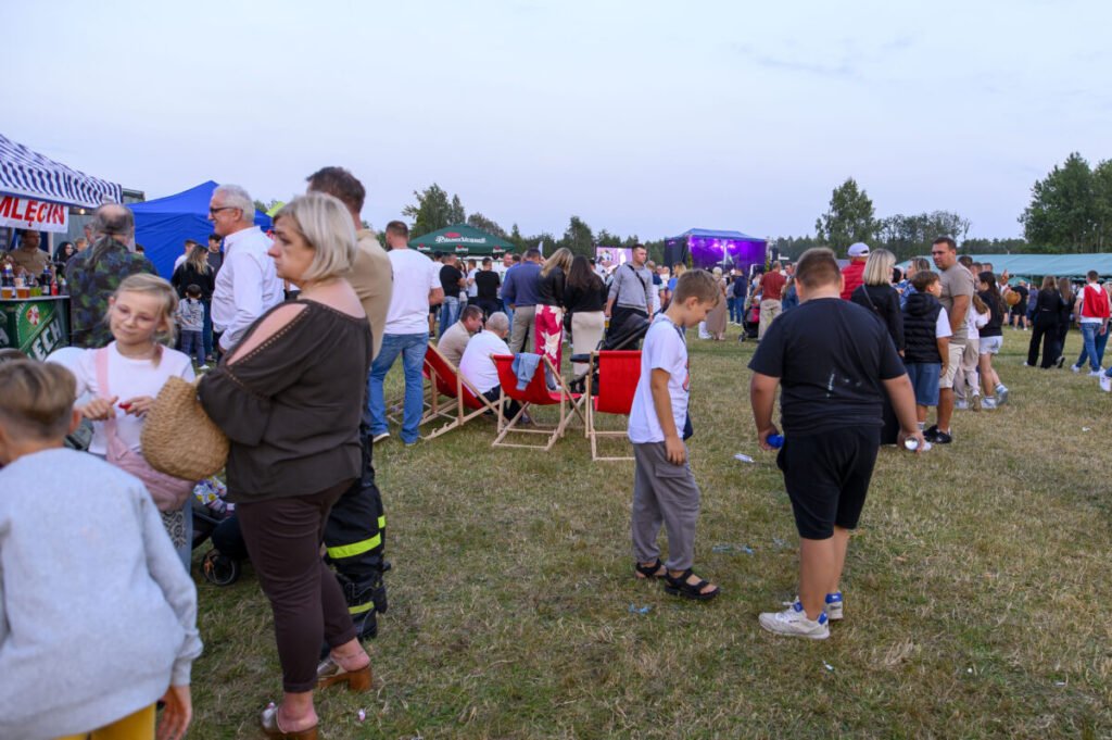Ludzie na festynie na trawie, liczne namioty i scena z kolorowymi światłami w tle. Dominują zielone, niebieskie i czerwone barwy. Atmosfera jest żywa i radosna.
