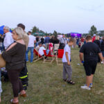 Ludzie na festynie na trawie, liczne namioty i scena z kolorowymi światłami w tle. Dominują zielone, niebieskie i czerwone barwy. Atmosfera jest żywa i radosna.