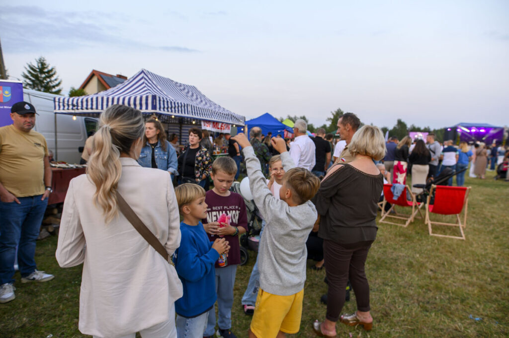 Grupa ludzi na festynie na tle stoiska z biało-niebieskim zadaszeniem. Trawiaste podłoże, w tle sceny z kolorowymi światłami. Dzień jest pogodny, niebo jasne.