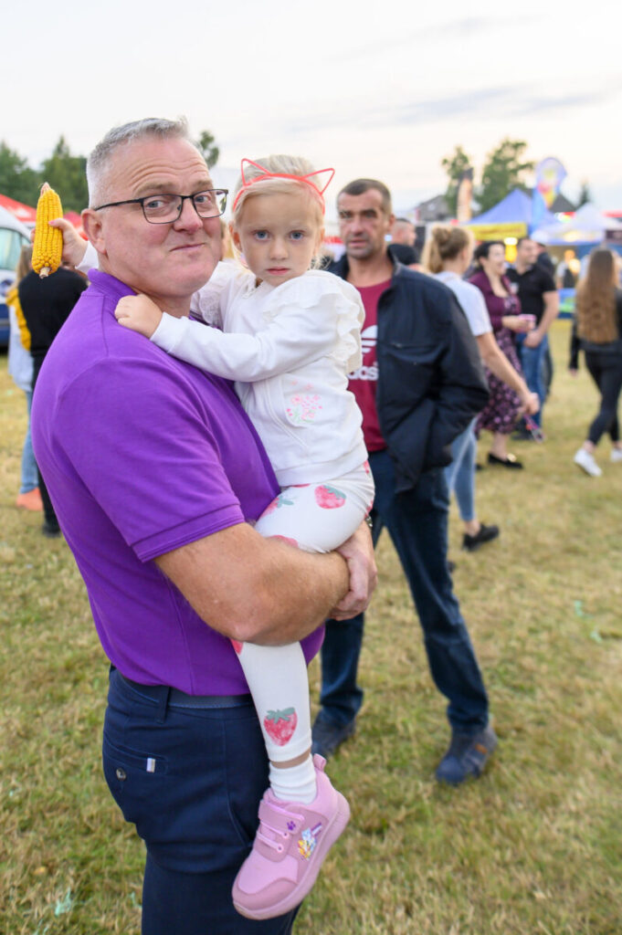 Mężczyzna w fioletowej koszulce trzyma dziecko na ramionach. Dziewczynka nosi różowe opaski na uszy. W tle plenerowe wydarzenie.