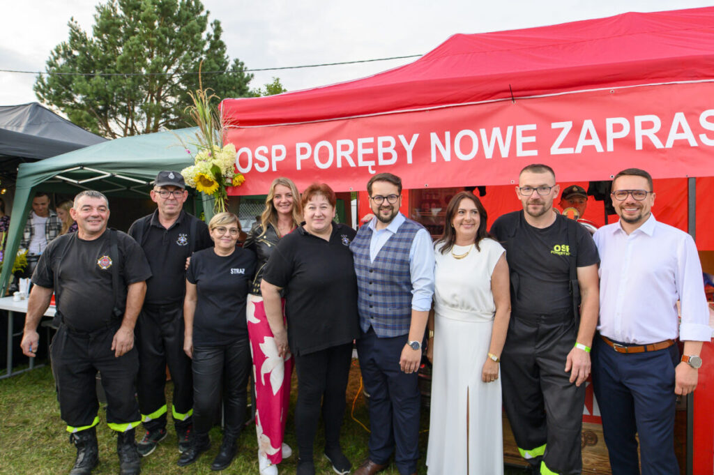 Grupa ludzi stoi przed czerwonym namiotem OSP, z zielonymi namiotami i drzewami w tle. Noszą czarne ubrania i białe stroje. Napis na namiocie to OSP Poręby Nowe zaprasza.