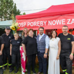 Grupa ludzi stoi przed czerwonym namiotem OSP, z zielonymi namiotami i drzewami w tle. Noszą czarne ubrania i białe stroje. Napis na namiocie to OSP Poręby Nowe zaprasza.