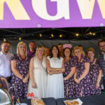 Grupa ludzi pozuje pod zadaszeniem z fioletowym banerem. Kobiety w kwiecistych sukienkach, mężczyźni w eleganckich koszulach. Na stole przed nimi widać jedzenie i dekoracje.