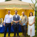 Obraz przedstawia cztery osoby stojące przed namiotem w kolorze żółtym z napisem Miód z rodzinnej pasieki. Osoby są elegancko ubrane, a tło stanowi trawa i drzewo. Przy namiocie widoczne są slój i plakaty związane z