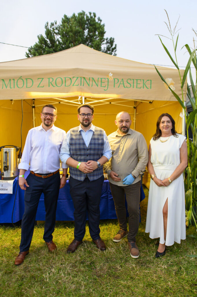 Stojąca kobieta w białym fartuchu mówi do pań siedzących przy stole, na którym stoją lusterka i leżą białe kartki papieru. W tle regały z książkami