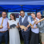 Grupa ludzi w namiocie trzyma drewniane rzeźby. Tło jest niebieskie z obrazami. Wszyscy się uśmiechają.