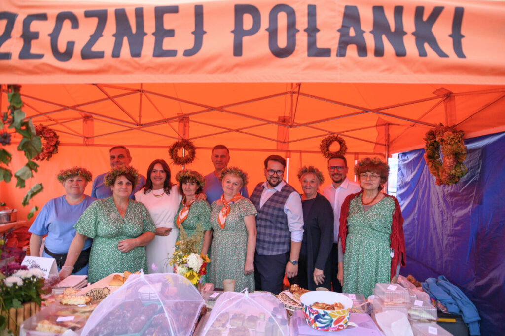 Grupa ludzi w strojach ludowych stoi pod pomarańczowym namiotem. Stoisko z jedzeniem i wieńcami z tyłu. Tło jest jednolite.