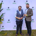 Dwóch mężczyzn stoi na tle ścianki z logo i napisem Gmina Dobre. Tło jest białe z zielonym i czarnym logo. Po bokach rośliny z żółtymi kwiatami.