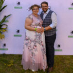 Para pozuje na tle białego baneru z napisem Gmina Dobre. Kobieta nosi różową suknię z kwiatowym wzorem, mężczyzna ma na sobie kamizelkę w kratę. Oboje stoją na trawie, obok zielonych roślin.