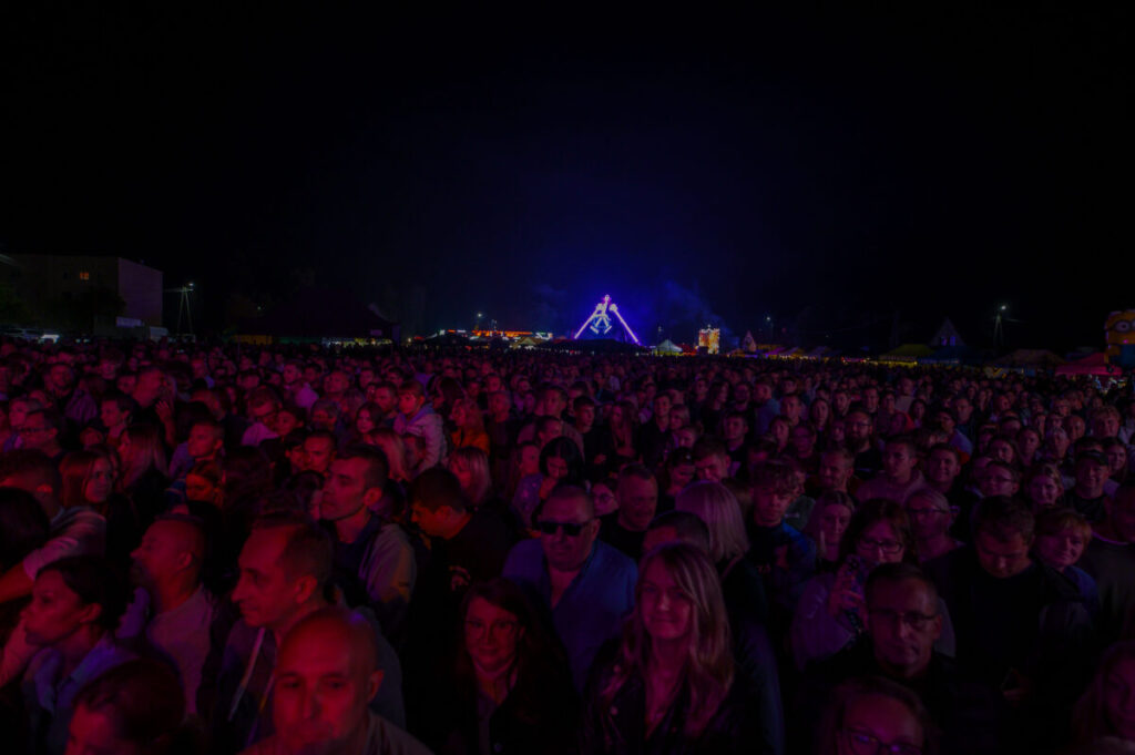 Tłum ludzi na koncercie nocą, oświetlony fioletowymi i niebieskimi światłami. W tle widać jasne, geometryczne dekoracje. Atmosfera wydarzenia jest żywa i pełna energii.