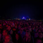 Tłum ludzi na koncercie nocą, oświetlony fioletowymi i niebieskimi światłami. W tle widać jasne, geometryczne dekoracje. Atmosfera wydarzenia jest żywa i pełna energii.