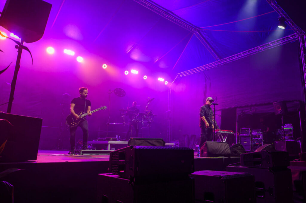 Zespół muzyczny na scenie pod fioletowym oświetleniem. Muzyk gra na gitarze, a inny śpiewa przy mikrofonie. W tle perkusista, sprzęt muzyczny i półprzezroczysty namiot.