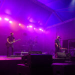 Zespół muzyczny na scenie pod fioletowym oświetleniem. Muzyk gra na gitarze, a inny śpiewa przy mikrofonie. W tle perkusista, sprzęt muzyczny i półprzezroczysty namiot.