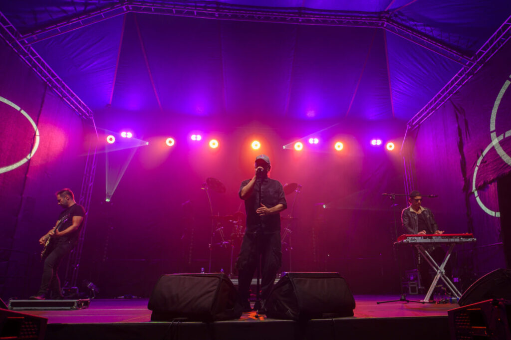 Zespół występuje na scenie oświetlonej fioletowym i pomarańczowym światłem. Na przodzie trzech muzyków: gitarzysta, wokalista, klawiszowiec. Tło to ciemny teatr pod namiotem.