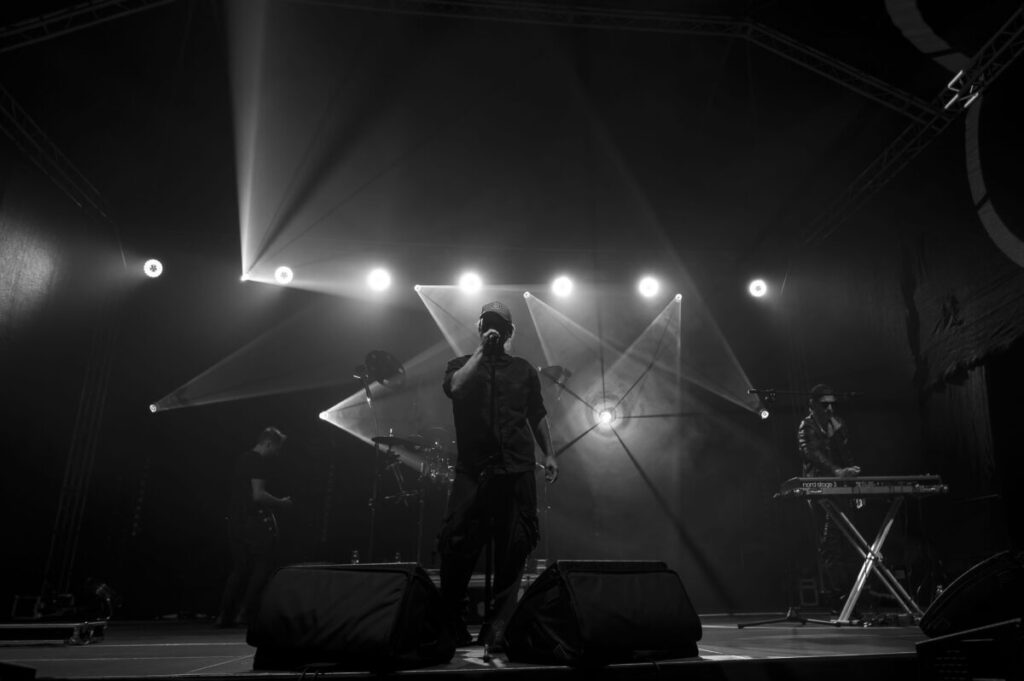Koncert na scenie, centralnie wokalista w czarnej koszuli. Jasne reflektory w tle tworzą dramatyczne oświetlenie. Klawiszowiec po prawej stronie.