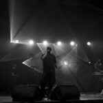 Koncert na scenie, centralnie wokalista w czarnej koszuli. Jasne reflektory w tle tworzą dramatyczne oświetlenie. Klawiszowiec po prawej stronie.