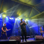 Koncert na scenie z kolorowymi reflektorami. Muzycy grają na gitarze i keyboardzie. Ciemne tło z jaskrawym oświetleniem.