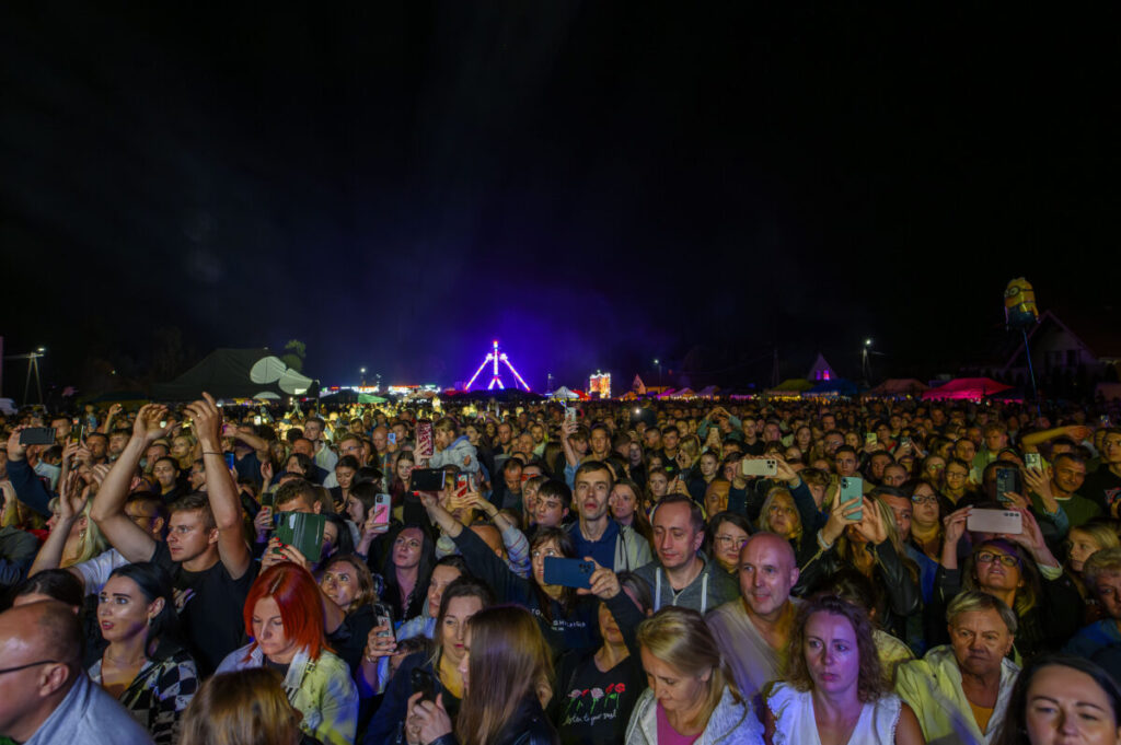 Tłum na koncercie w nocy; kolorowe światła w tle, ciemne niebo. Ludzie robią zdjęcia telefonami.