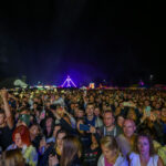 Tłum na koncercie w nocy; kolorowe światła w tle, ciemne niebo. Ludzie robią zdjęcia telefonami.