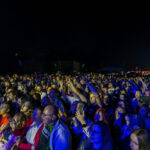 Tłum ludzi na koncercie nocą, oświetlony niebieskim światłem. W tle rozmyte światła lunaparku. Radosna atmosfera.