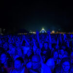 Tłum zgromadzony na koncercie nocą, oświetlony niebieskim światłem. W tle widać kolorowe, luminujące dekoracje. Ludzie podnoszą ręce.