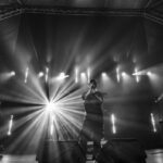 Trzech muzyków na scenie z jasnymi reflektorami w tle. Czarny i biały obraz podkreśla intensywność występu. Środkowy artysta trzyma mikrofon.