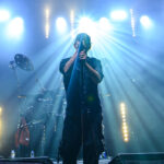 Osoba w czapce śpiewa na scenie z mikrofonem. Za nią jasne światła tworzą promienie. W tle perkusja w zimnym błękicie.