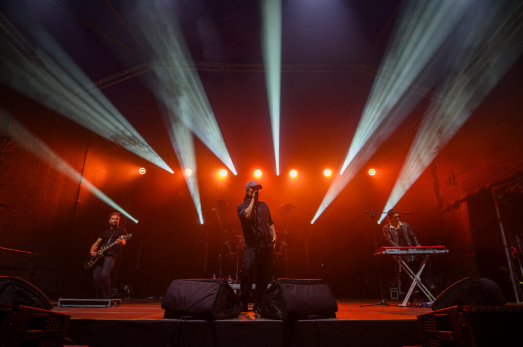 Koncert na scenie z czerwonymi światłami. Trzech muzyków, w tym wokalista w czapce, stoją na scenie. Tło jest ciemne.