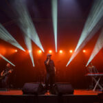 Koncert na scenie z czerwonymi światłami. Trzech muzyków, w tym wokalista w czapce, stoją na scenie. Tło jest ciemne.