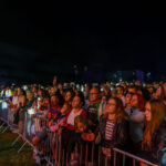 Na zdjęciu tłum ludzi stoi przed barierkami podczas nocnego koncertu. Oświetlenie sceniczne tworzy kolorowe refleksy na twarzach. W tle widoczne ciemne niebo i delikatne światła.