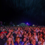 Tłum zgromadzony na koncercie nocą, oświetlony kolorowymi światłami. Tło ciemne z widocznymi sylwetkami. Atmosfera radosna.