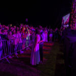 Tłum ludzi za barierkami na koncercie w kolorowym oświetleniu. Na scenie ekran z projekcją. Tło ciemne, wydarzenie nocne.