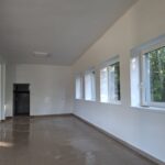 Pusty pokój z białymi ścianami i dużymi oknami po prawej stronie. Na podłodze jasne płytki. W tle drzwi wejściowe.