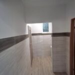 Korytarz z jasnobeżowymi płytkami i drewnianymi akcentami. Białe ściany i sufit, okno na końcu. Drzwi po lewej stronie.