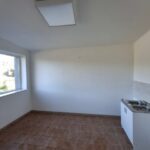 Pusta kuchnia z białymi ścianami i brązowymi płytkami na podłodze. Prosty zlew z szafką po prawej stronie. Duże okno, wpuszczające światło.